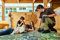 Bild vergrößert sich per Mausklick: Drei Jugendliche sitzen und liegen auf einem Fototeppich, der ein Luftbild der Umgebung zeigt. Ein Mitarbeiter des Forstmuseums kniet daneben und gibt Erklärungen.