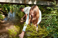 Bild vergrößert sich per Mausklick: Ein Mädchen und ein Junge stehen gebeugt in einem Bach und sind auf der Suche nachkleinen Wasserbewohnern